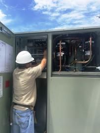 A technician wearing a hard hat performs maintenance inside an open, cabinet-style industrial HVAC unit outdoors.