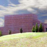 A large, multi-level brick building sits behind a grassy hill under a bright, cloudy sky with small evergreen trees.