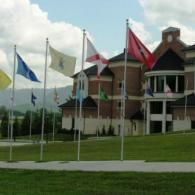 Several flags wave on poles in front of a multi-story brick building with a rounded entrance under a cloudy blue sky.