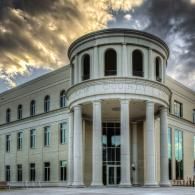 A grand neoclassical building with a circular pillared entrance under a dramatic, cloudy sunset sky.