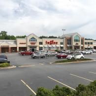 An Ingles grocery store and adjacent retail shops in a strip mall with a parking lot and parked cars under a cloudy sky.