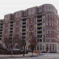 A multistory red brick apartment building with curved corner balconies, situated on a corner street with trees in front.