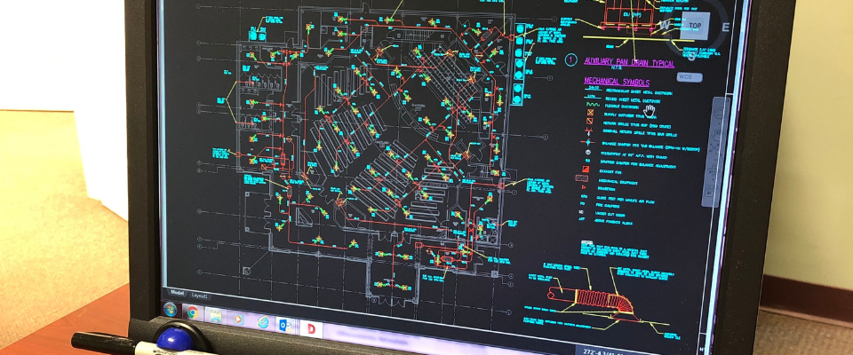 A computer monitor displays a complex CAD floor plan with various colored lines and technical symbols on a black background.