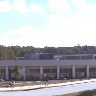 A low-slung, modern gray commercial building with large glass windows, surrounded by trees under a bright blue sky.