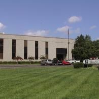 A beige two-story commercial building with dark windows sits behind a large mowed lawn under a clear blue sky.