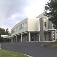 Modern two-story building with a curved glass facade, stone accents, and covered entrances under a cloudy sky.