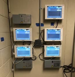 Five industrial touchscreen display units mounted on a beige wall with associated power boxes and electrical conduits.