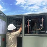 A person wearing a white hard hat and tan shirt services the electrical controls inside an industrial HVAC unit.