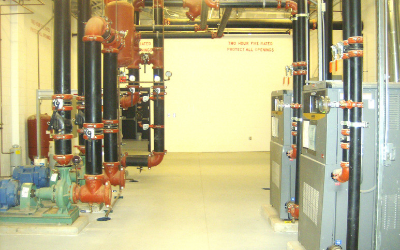 Mechanical room featuring industrial black pipes with orange connectors and large gray boiler units on a concrete floor.