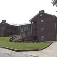 A multi-story, dark brick apartment building with a grassy lawn, bushes, and an outdoor staircase in front.