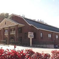 A two-story brick building with a columned front portico and a sign stand on a sunny day with red shrubs in the foreground.