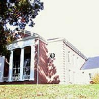 A brick-and-white academic building with white columns, set on a lawn with a large tree in the foreground.