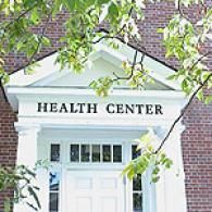 White entrance to a brick health center building with a pediment roof and surrounding green tree branches.