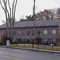 A two-story brick building with rows of windows and a gray roof, surrounded by a metal fence and tall bare trees.