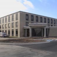 A multi-story beige building with a covered entrance portico, featuring rows of windows and a parked van under a blue sky.