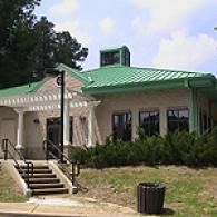 A single-story brick building with a green metal roof, a white pergola entryway, and concrete steps leading to the door.