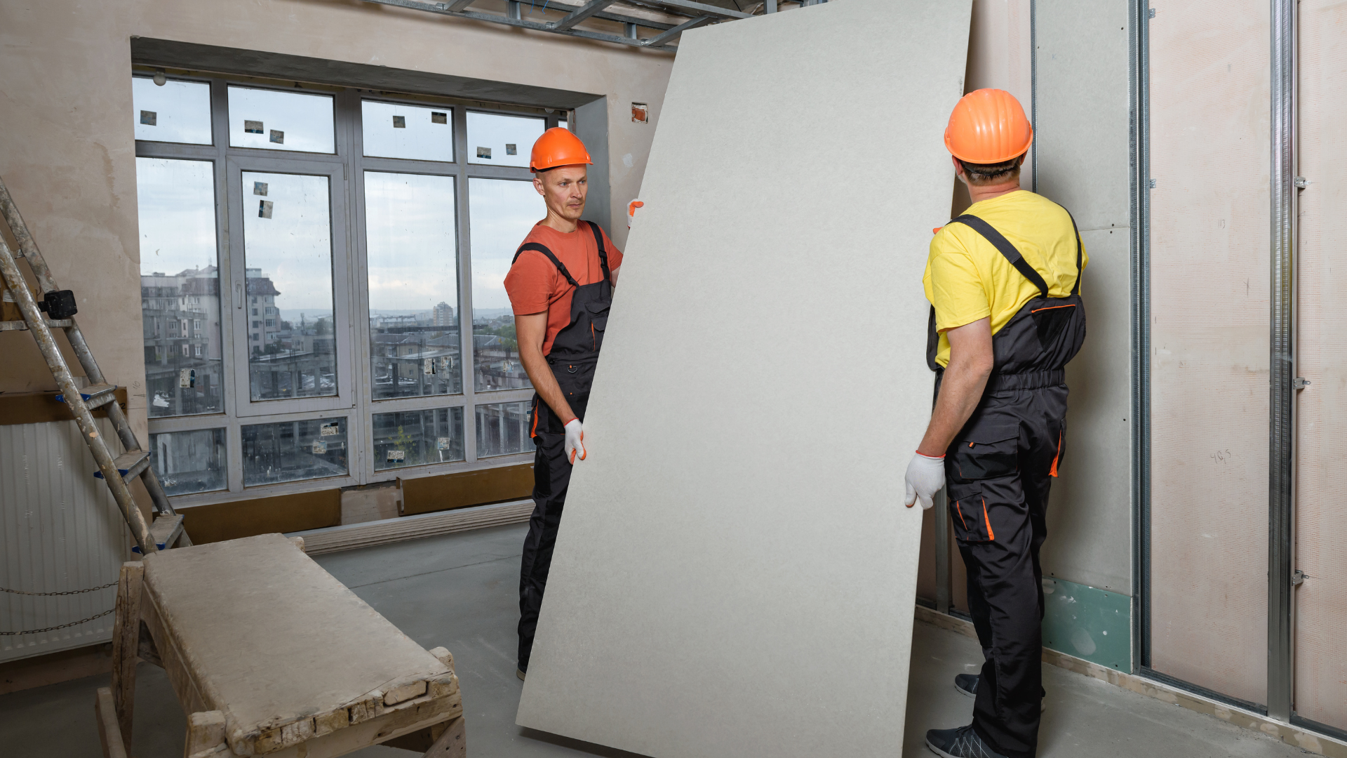 Two construction workers are carrying a large piece of drywall in a room.