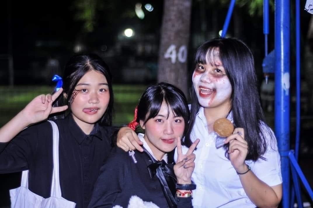 Three young people pose together at night; two smile while making peace signs, and one wears face paint resembling a spooky Halloween look while holding a cookie.