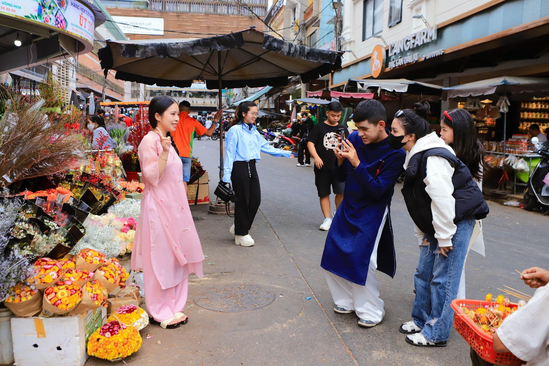 A young Vietnamese woman in a pink ao dai posing for a photo at a vibrant flower market during Tet holiday.