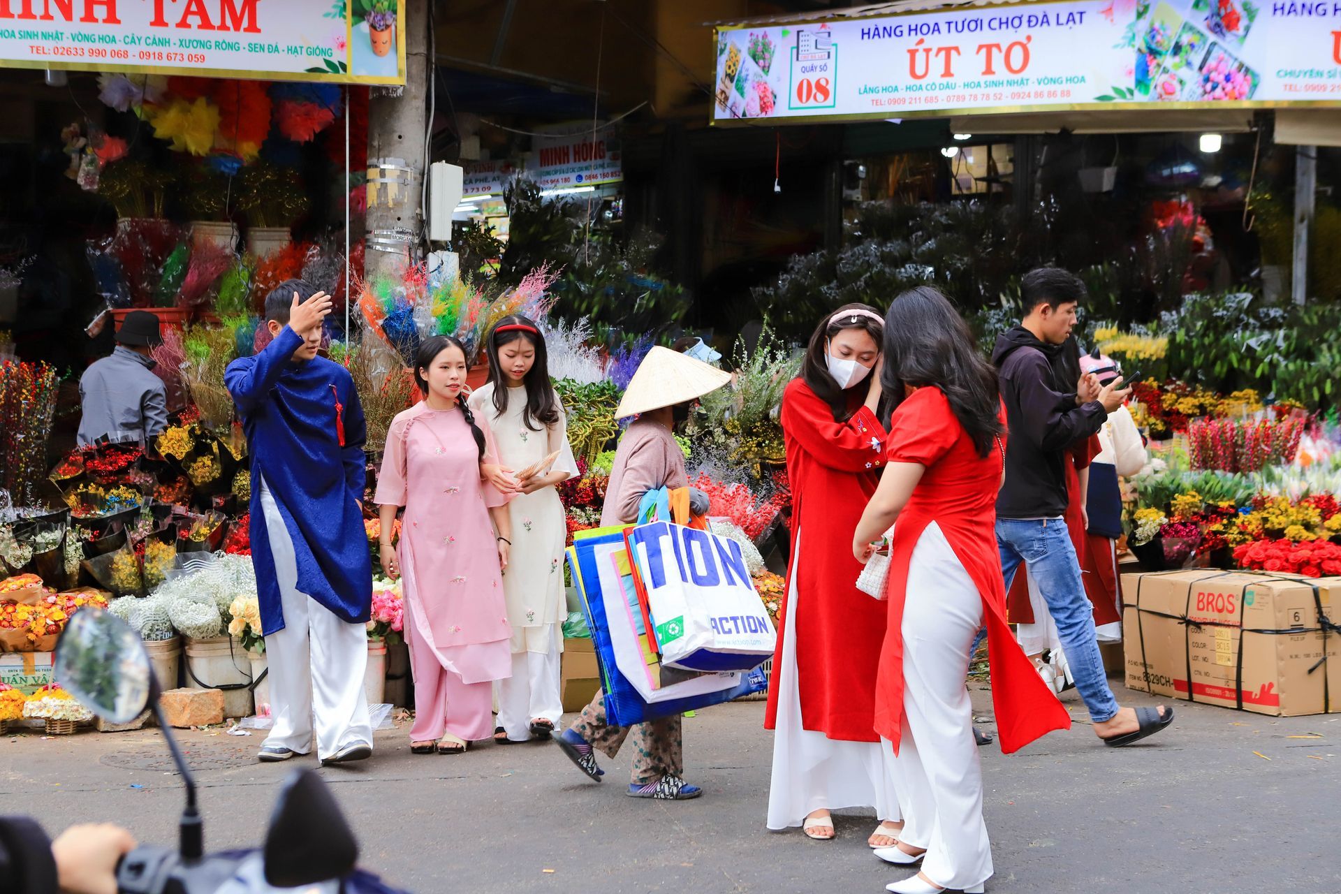 A group of Vietnamese women and children in traditional ao dai taking photos surrounded by colorful flowers at a Tet market.