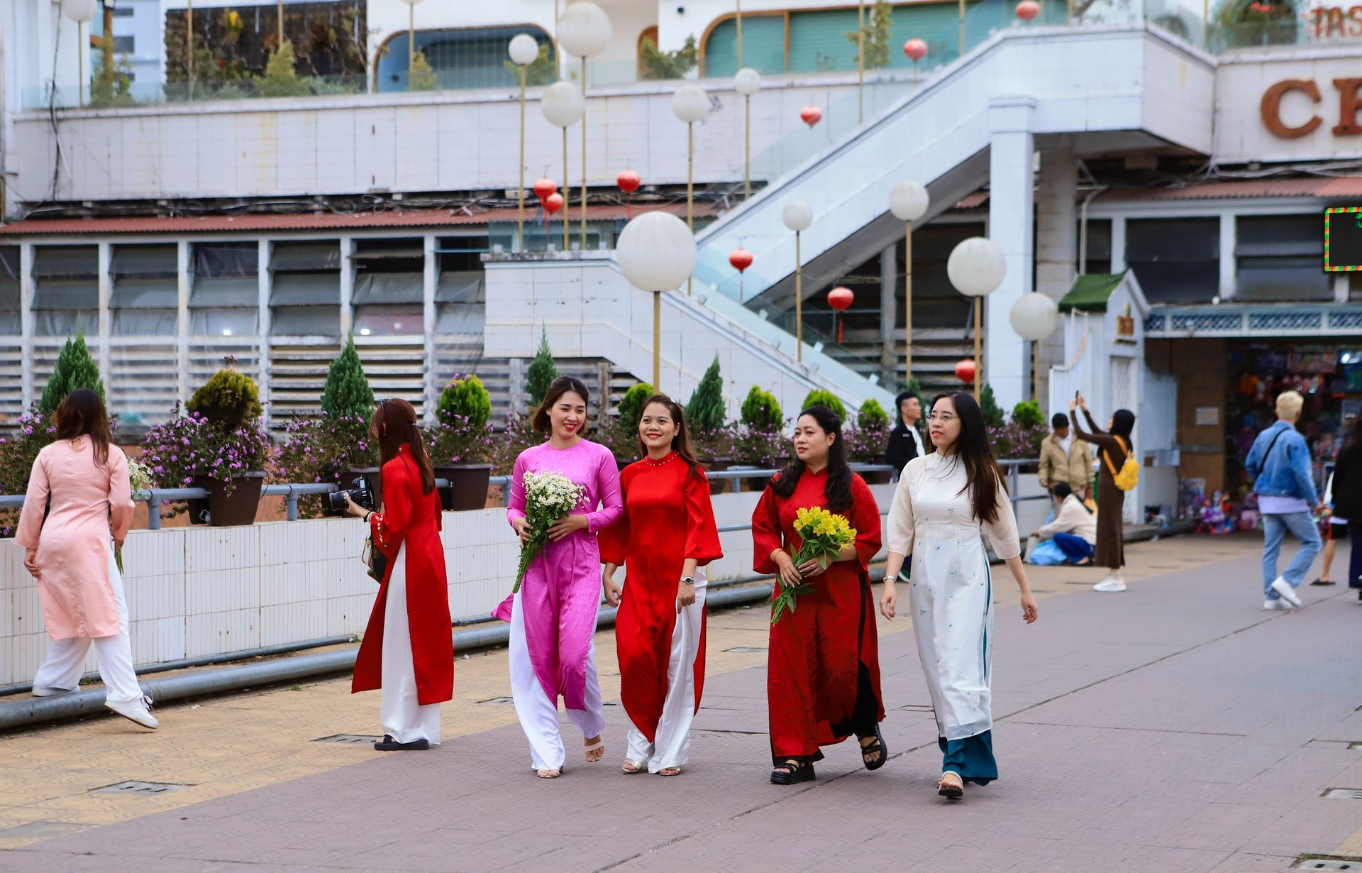 A group of young Vietnamese women in colorful ao dai walking together, holding flowers, and enjoying Tet festivities in an urban setting.