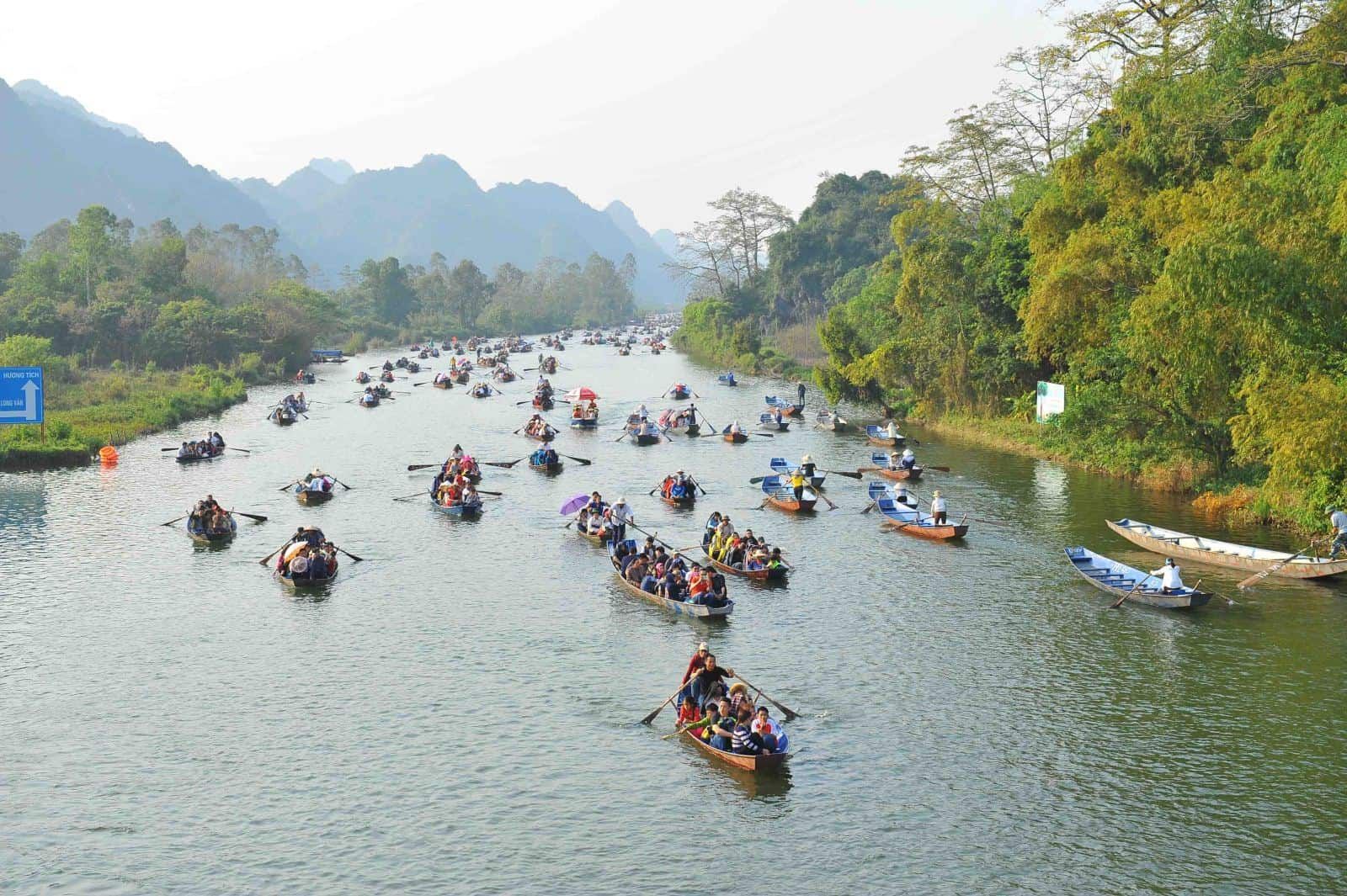 Tourists riding traditional wooden boats along a river surrounded by green mountains, heading toward the Perfume Pagoda in Vietnam.