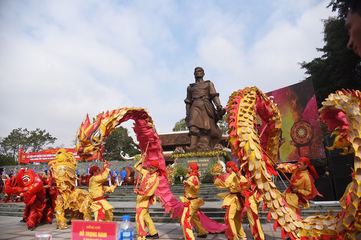 A dragon dance performance with vibrant red and yellow dragons in front of the King Quang Trung statue during a Vietnamese spring festival.