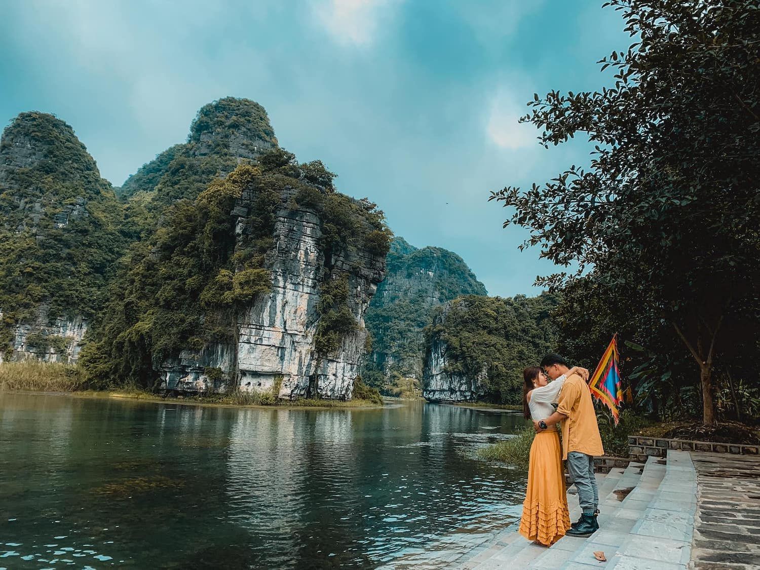 A couple in traditional Vietnamese ao dai standing by a calm river, surrounded by limestone karst mountains in Ninh Binh, Vietnam.