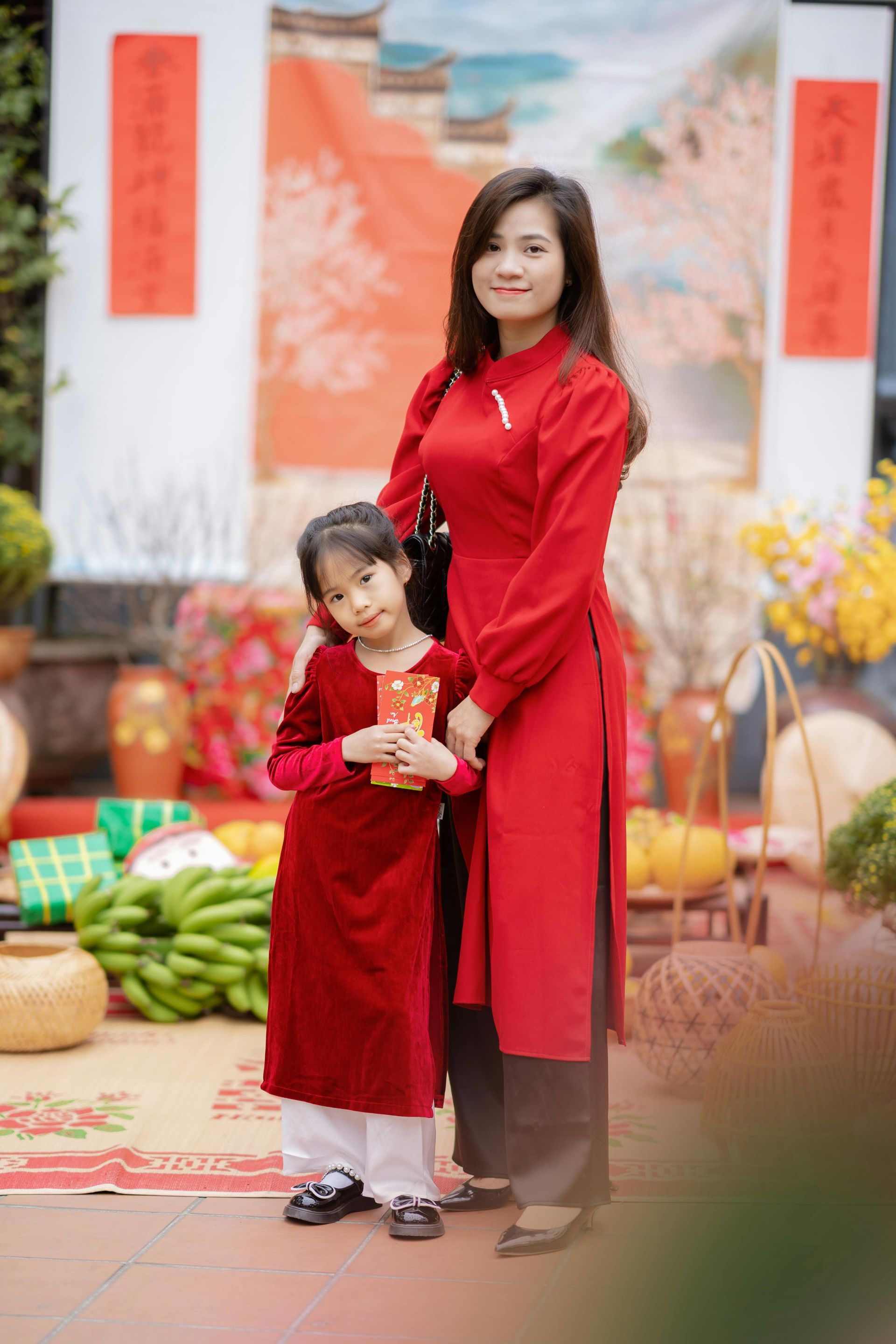 A young Vietnamese girl in a red ao dai holding a red envelope (li xi) while standing close to her mother during Tet holiday.