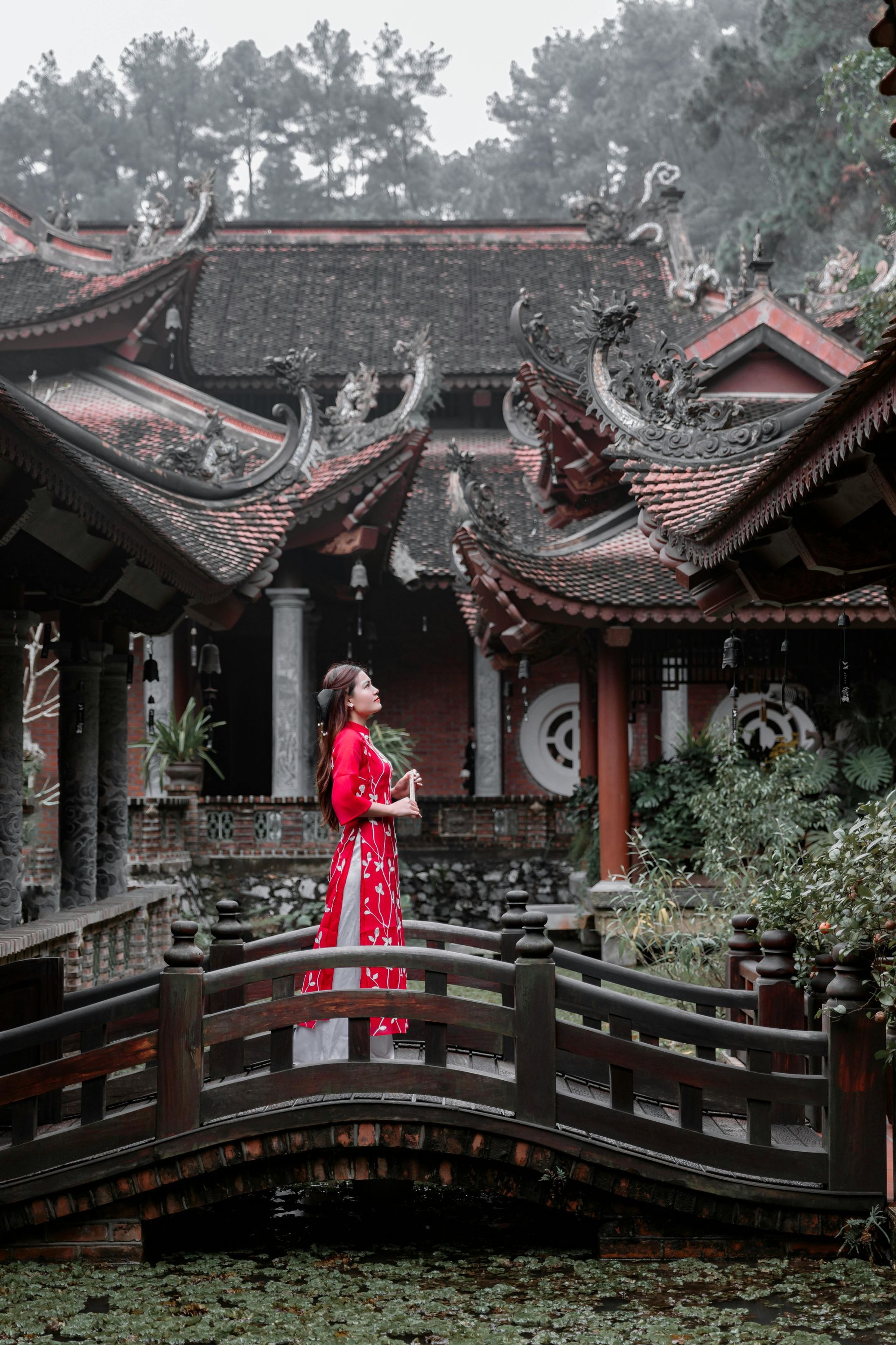 A woman in a red áo dài standing beside an ancient Vietnamese temple with curved roofs and intricate wooden carvings.