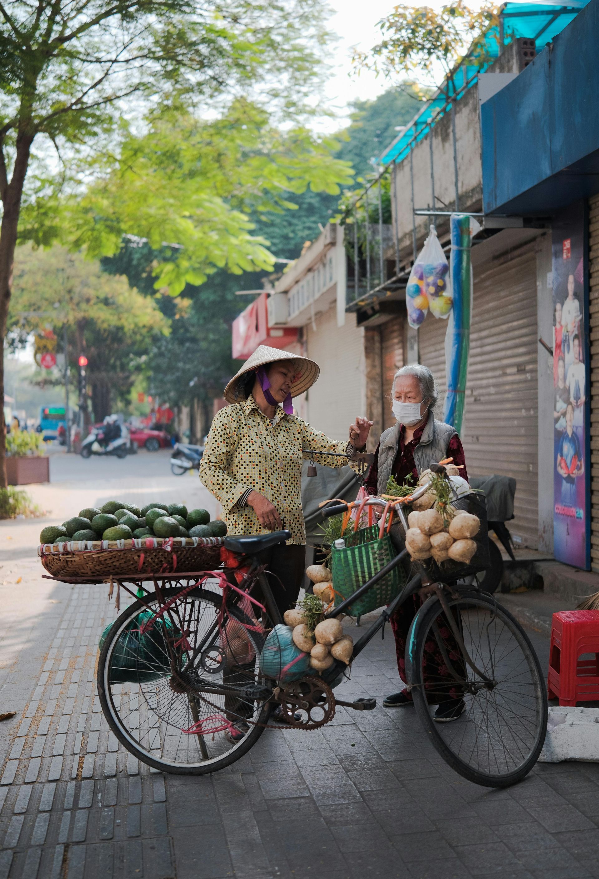 Two Vietnamese women with bicycles selling fresh fruits such as avocados and coconuts on a sidewalk, engaging in conversation