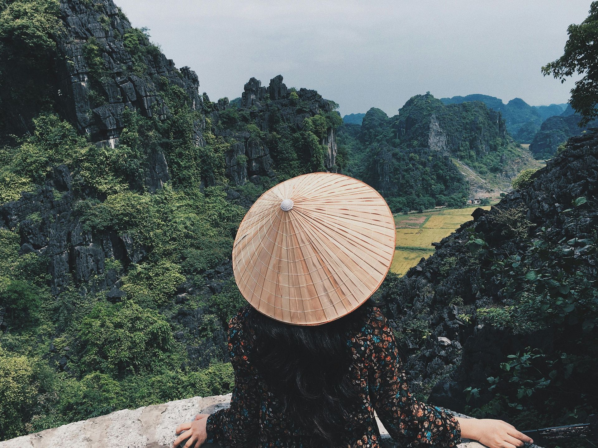 A woman wearing a conical hat sitting and gazing at the serene natural landscape below.