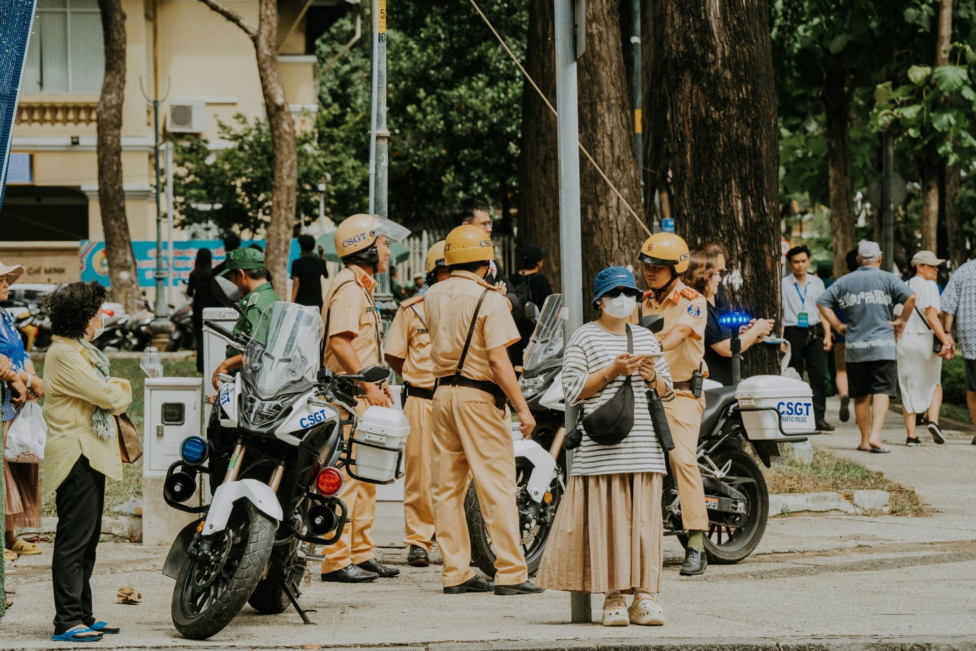 Vietnamese traffic police officers wearing yellow uniforms and helmets, standing beside their motorcycles while interacting with pedestrians on a city street.