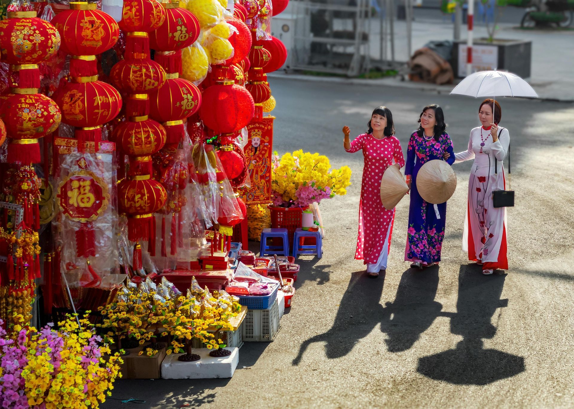 Three Vietnamese women in red and white ao dai walking past a display of red lanterns and yellow flowers, symbolizing Tet celebrations.