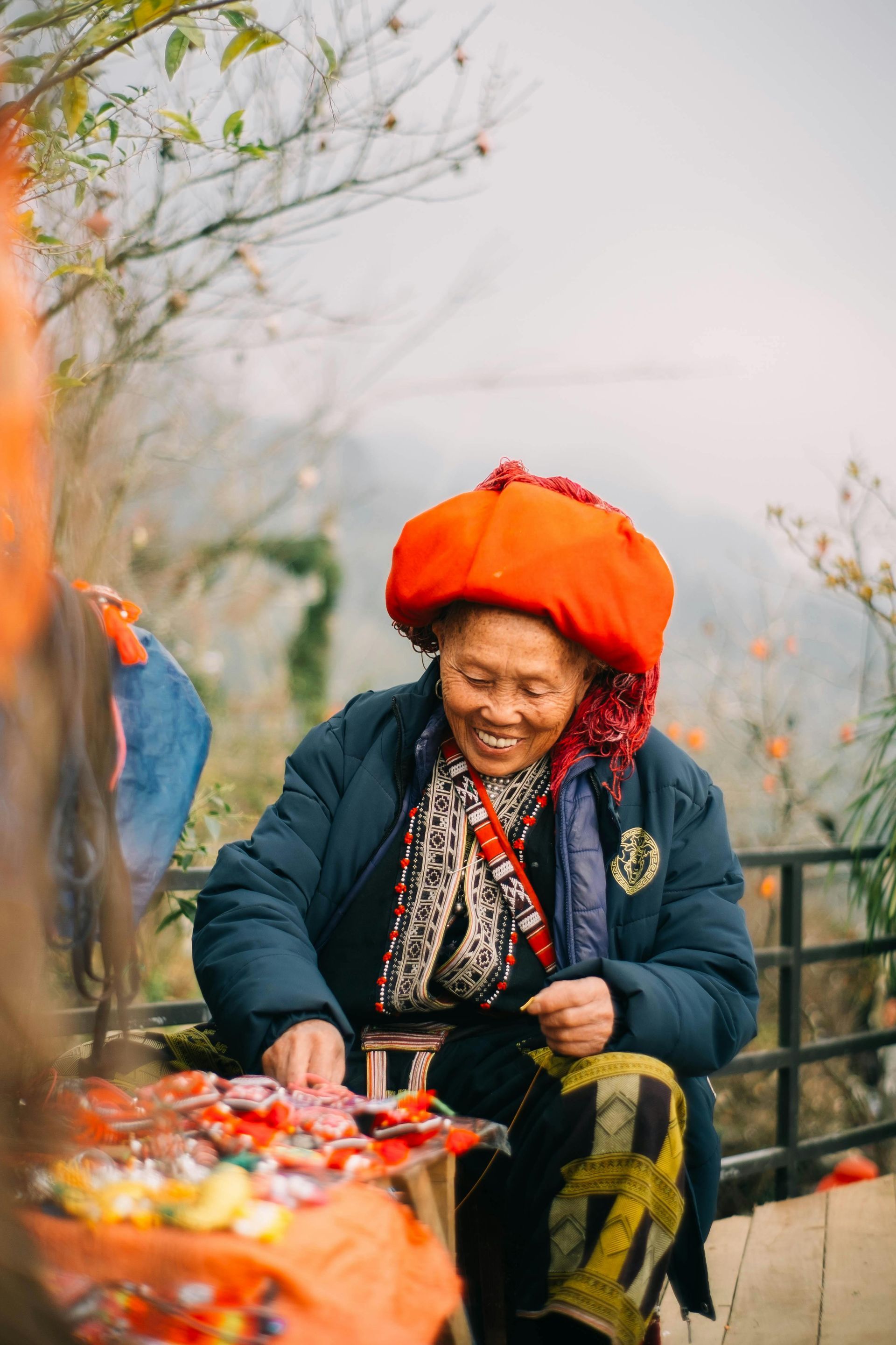 A smiling elderly woman from an ethnic minority group in northern Vietnam, wearing a traditional red headscarf and cultural attire.