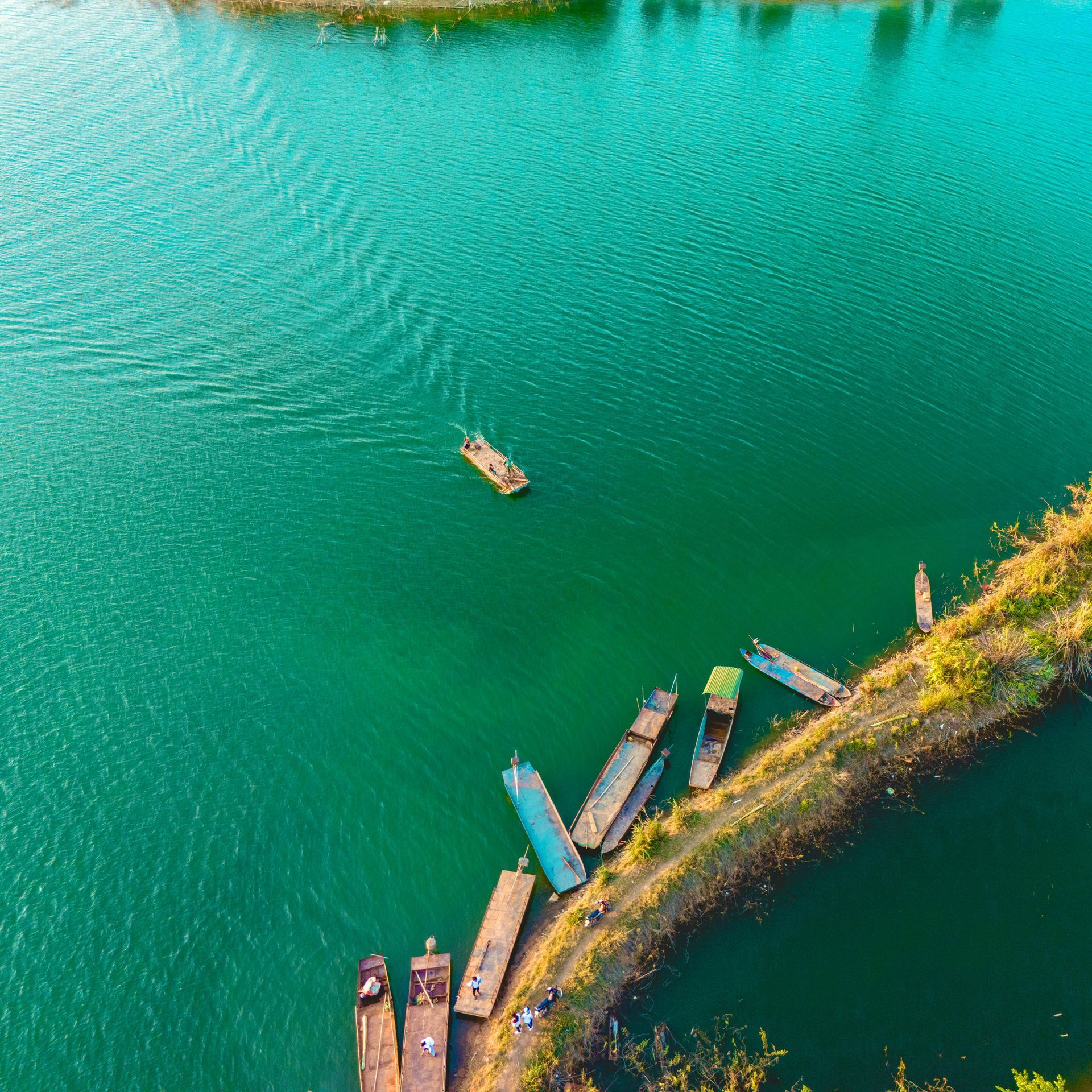 An aerial view of emerald-green waters with a small wooden boat and colorful traditional boats anchored along a shoreline in Vietnam.