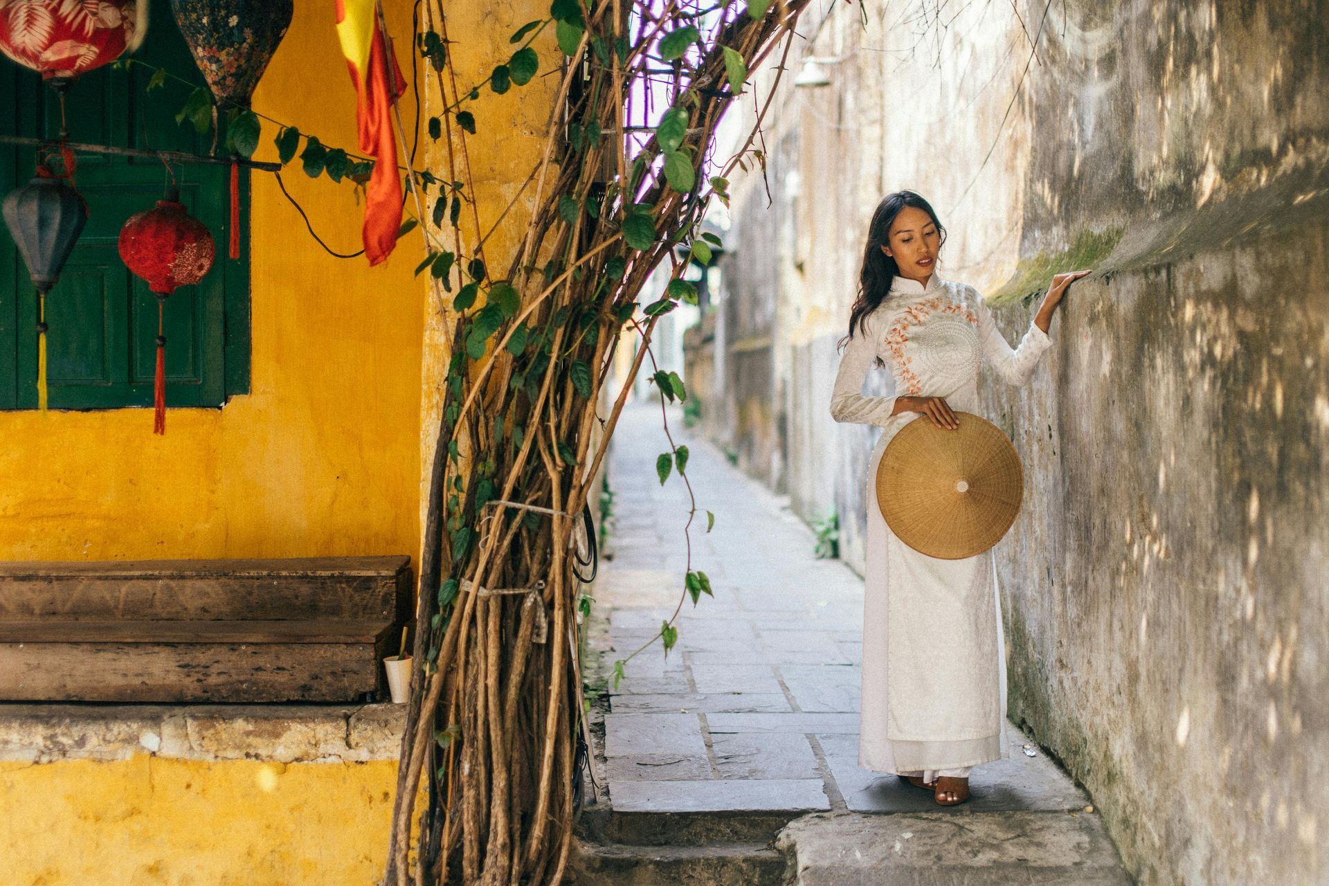 A woman in a white áo dài and nón lá walking in Hội An’s iconic yellow alley.