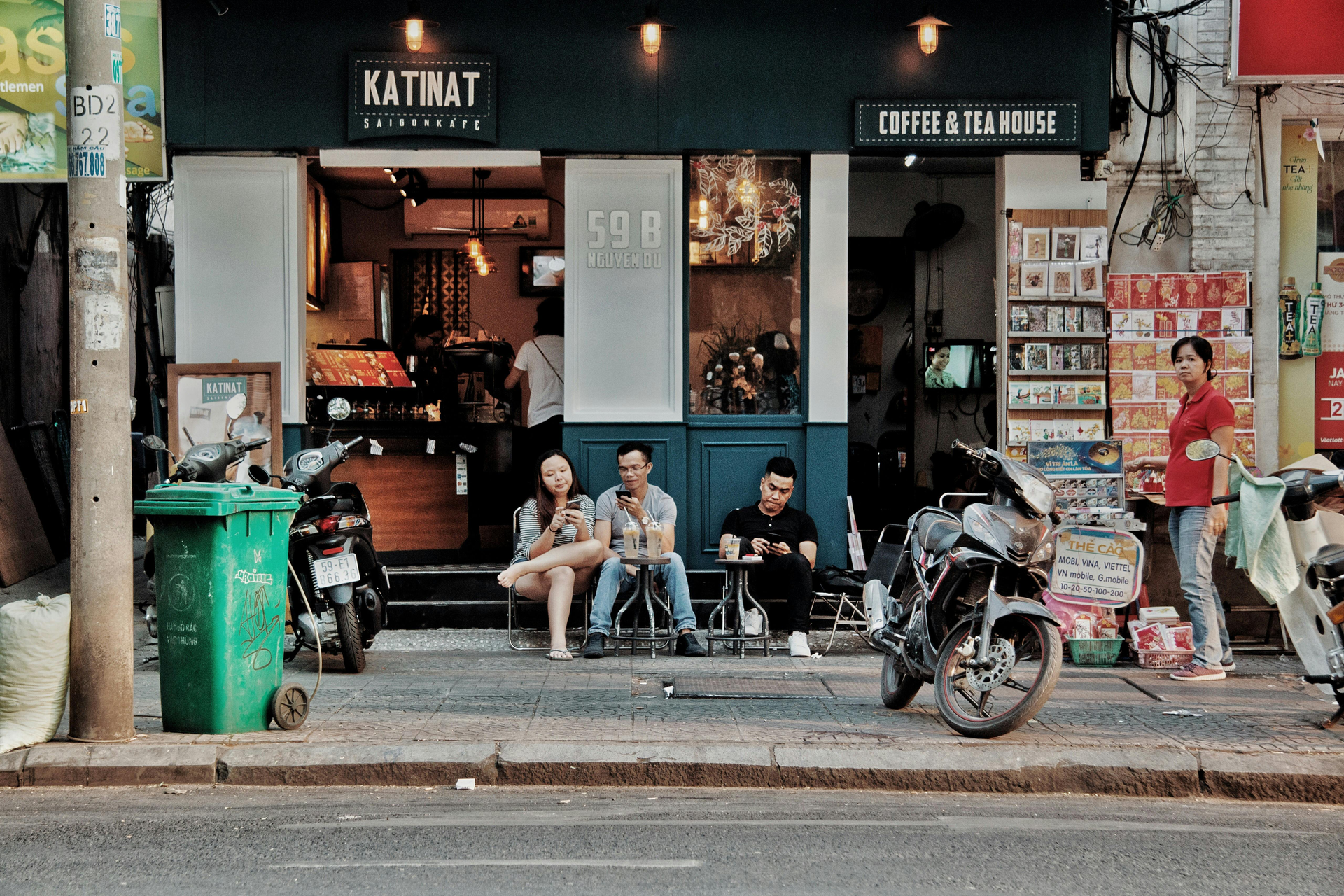 A street scene in Vietnam, showing people sitting outside a local coffee shop, motorcycles parked nearby, and a relaxed urban atmosphere.