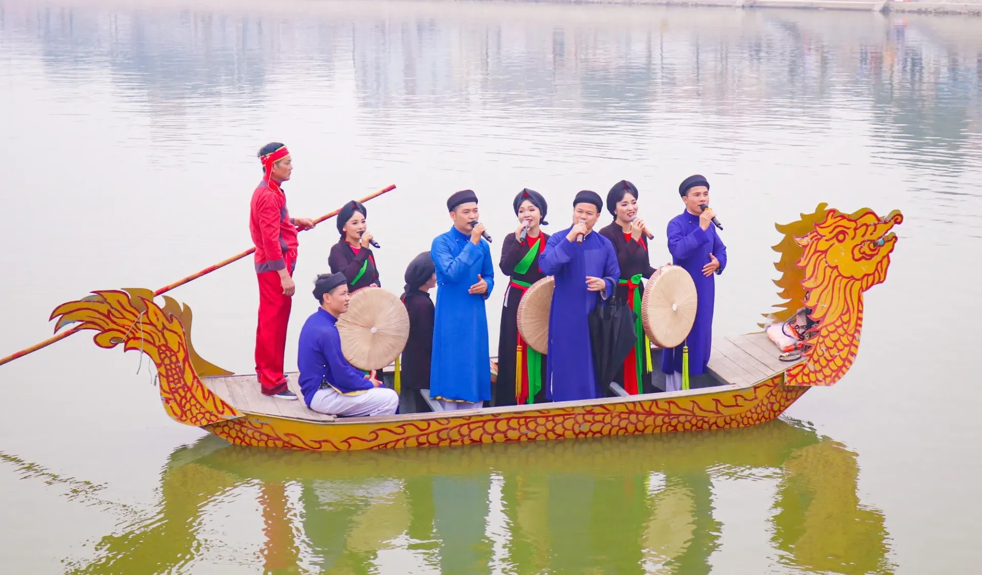 A group of men and women in traditional Vietnamese ao dai performing Quan Ho folk singing on a dragon boat floating on a calm river.