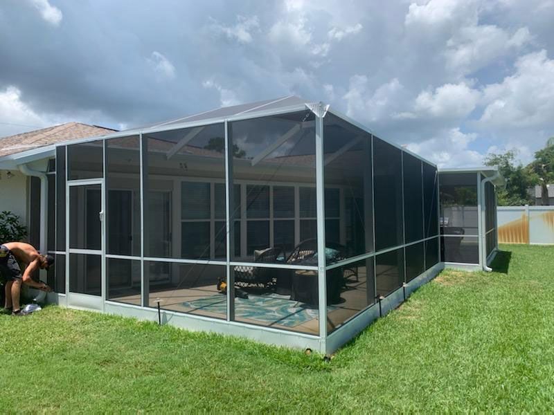 Screened patio enclosure beside a house with a table and chairs on a grassy lawn under cloudy skies