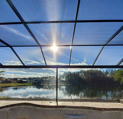 Sunlit lake view through a screened patio, with blue sky, trees, and calm water reflected below