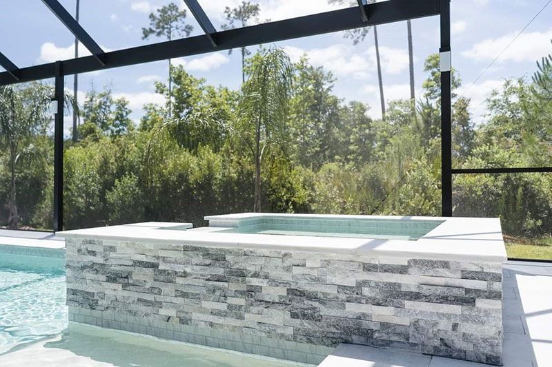 Modern outdoor hot tub beside a pool under a screened pergola, with stone-clad sides and trees beyond