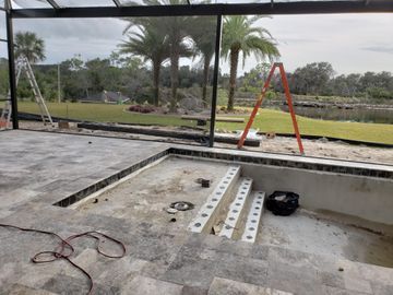 Partially built concrete pool with ladder, orange sawhorse, and palm trees by a grassy field