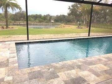 Covered backyard pool with stone patio and screen enclosure overlooking a grassy pond area