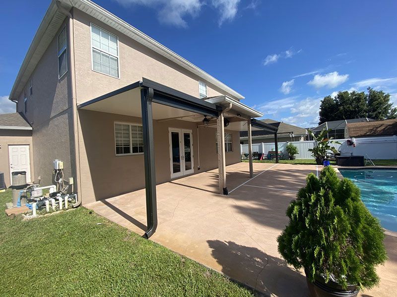 Back patio with tan house, covered pergola, concrete patio, and green lawn under blue sky