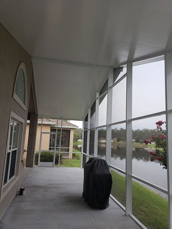 Covered patio with a grill beside glass windows, overlooking a grassy lakeside yard.