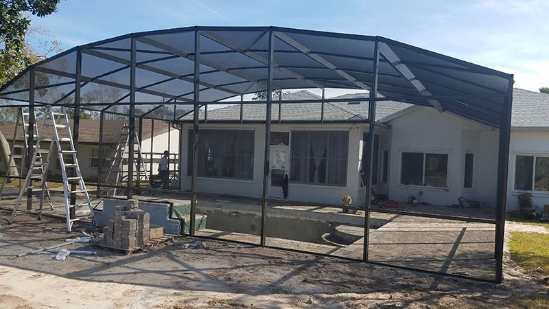 Metal carport frame under construction in front of a white building, with ladders and tools on site