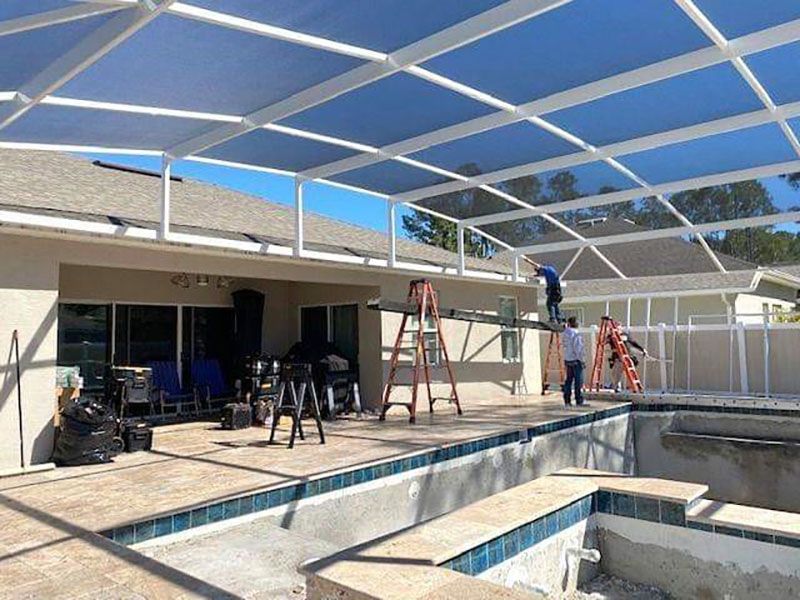Workers installing a blue shade canopy over an outdoor pool area with ladders and equipment.