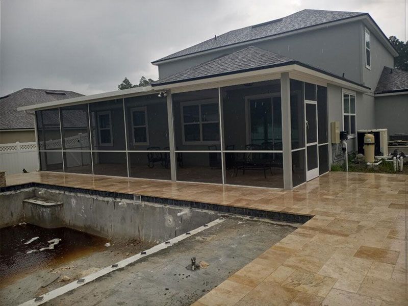 Modern house with screened patio and beige stone patio beside a partially filled pool.