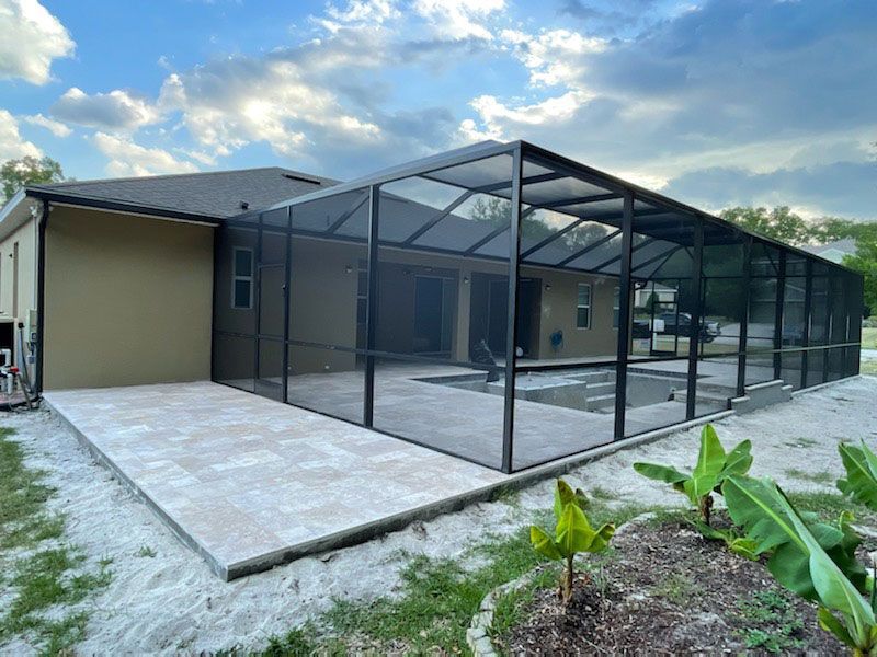 Screened patio enclosure beside a beige house with a tiled concrete patio and tropical plants.